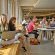 Studierende in der Aula der AAU. Vorne sitzt eine junge Studentin mit Laptop am Fenster in der Aula.