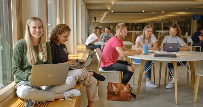 Studierende in der Aula der AAU. Vorne sitzt eine junge Studentin mit Laptop am Fenster in der Aula.