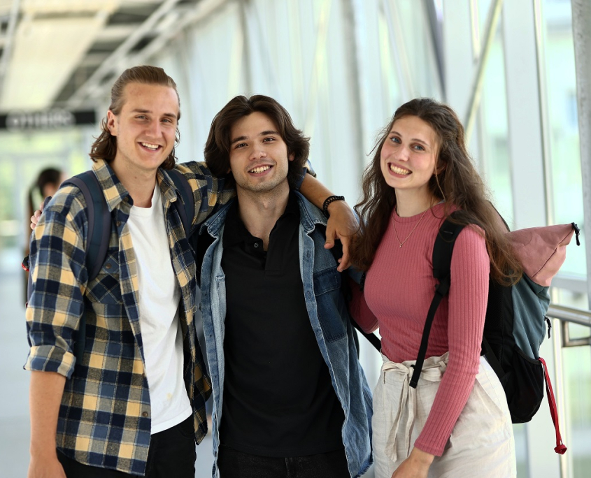 Drei Studierende stehen lachend in Verbindungsgang zwischen Zentralgebäude und Seitengebäude der Universität Klagenfurt.