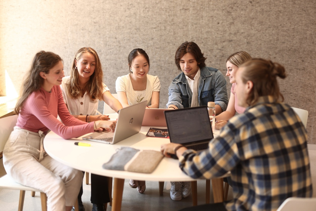 Eine Lerngruppe aus fünf Studierenden sitzt an einem runden Tisch vor der Universitätsbibliothek der Universität Klagenfurt und arbeitet an Laptops.