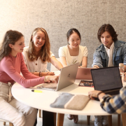 Eine Lerngruppe aus fünf Studierenden sitzt an einem runden Tisch vor der Universitätsbibliothek der Universität Klagenfurt und arbeitet an Laptops.