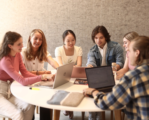 Eine Lerngruppe aus fünf Studierenden sitzt an einem runden Tisch vor der Universitätsbibliothek der Universität Klagenfurt und arbeitet an Laptops.