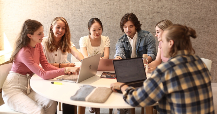 Eine Lerngruppe aus fünf Studierenden sitzt an einem runden Tisch vor der Universitätsbibliothek der Universität Klagenfurt und arbeitet an Laptops.