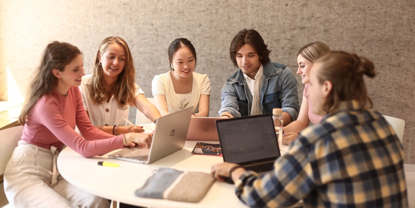 Eine Lerngruppe aus fünf Studierenden sitzt an einem runden Tisch vor der Universitätsbibliothek der Universität Klagenfurt und arbeitet an Laptops.