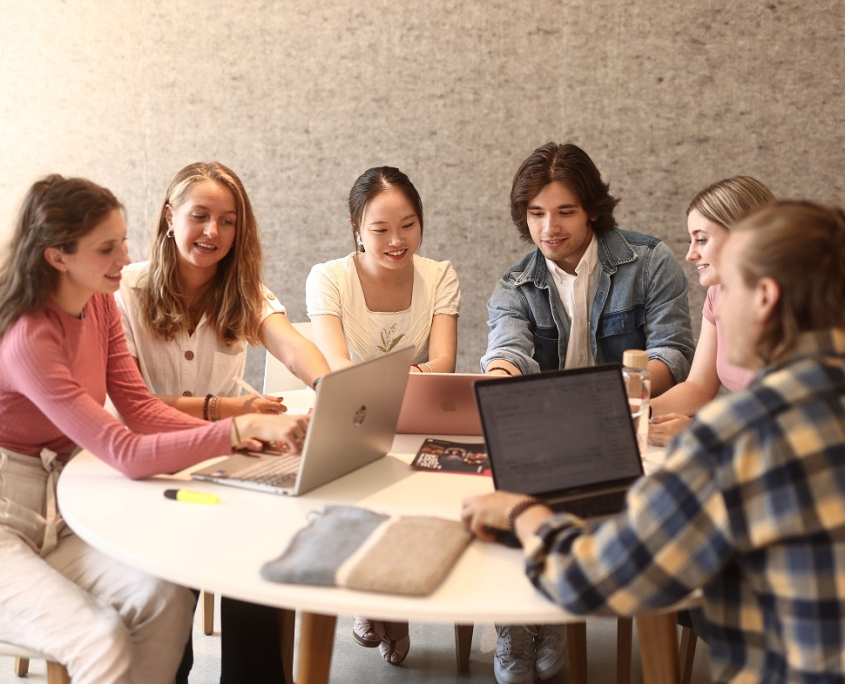 Eine Lerngruppe aus fünf Studierenden sitzt an einem runden Tisch vor der Universitätsbibliothek der Universität Klagenfurt und arbeitet an Laptops.
