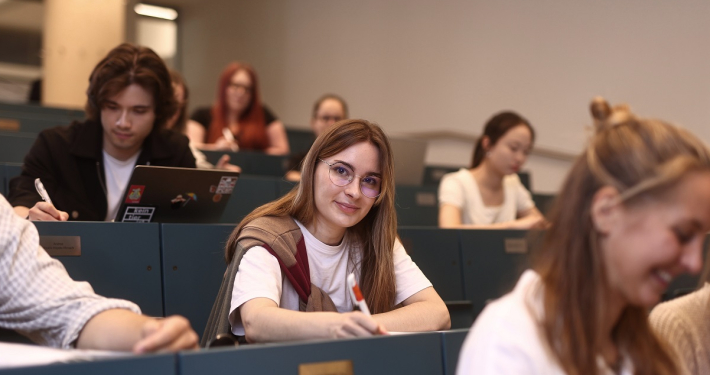 Mehrere Studierende sitzen in den gestuften blaufarbigen Sitzreihen eines Hörsaals.