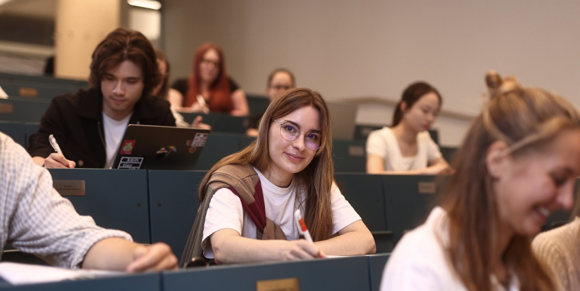 Mehrere Studierende sitzen in den gestuften blaufarbigen Sitzreihen eines Hörsaals.