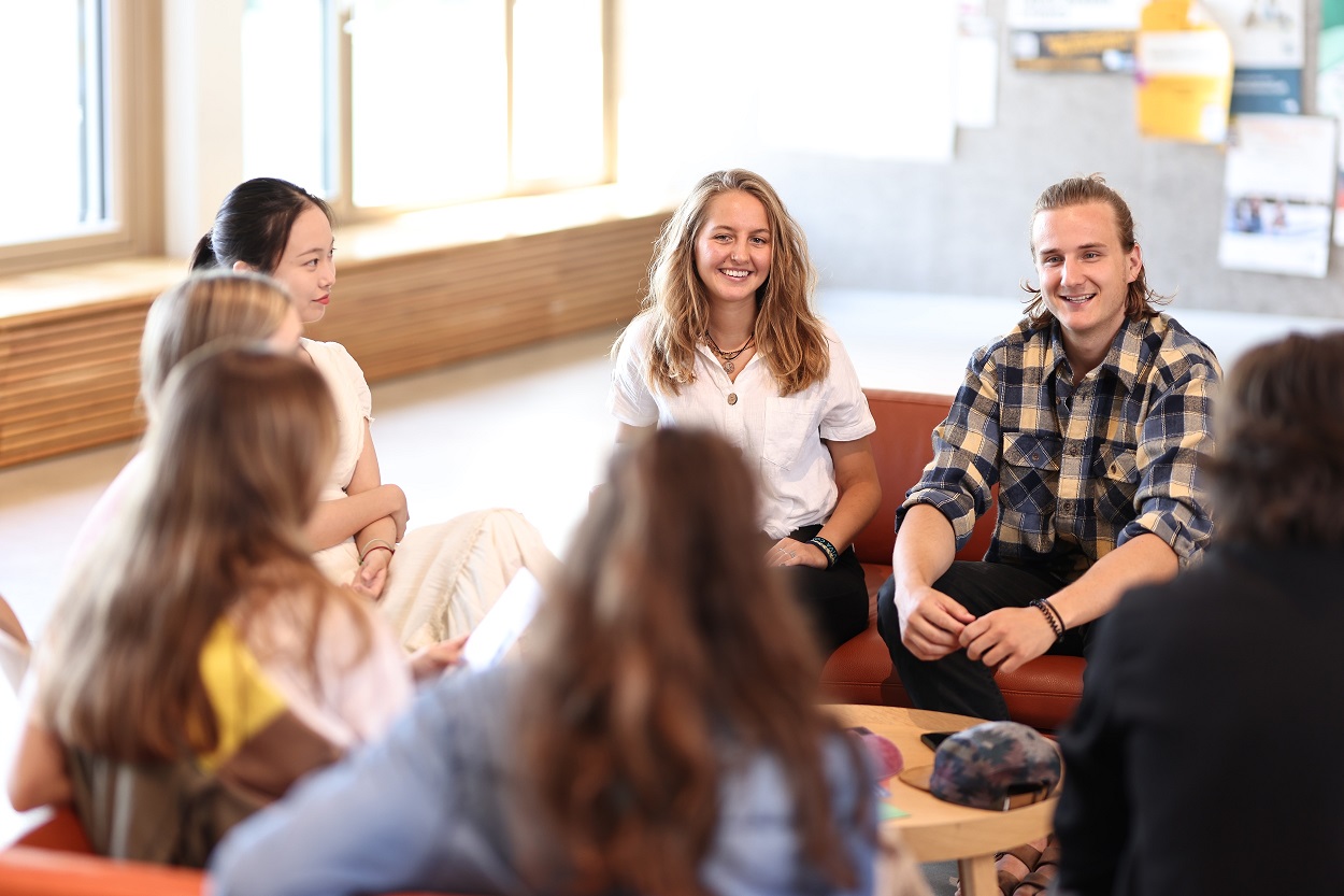 Studierende sitzen auf orangefarbenen Polstermöbeln in der Aula um einen runden niedrigen Holztisch.