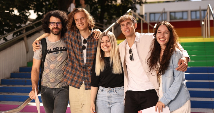 Eine Gruppe von fünf Studierenden steht lachend vor Treppe in Regenbogenfarben auf dem Campus der Universität Klagenfurt.