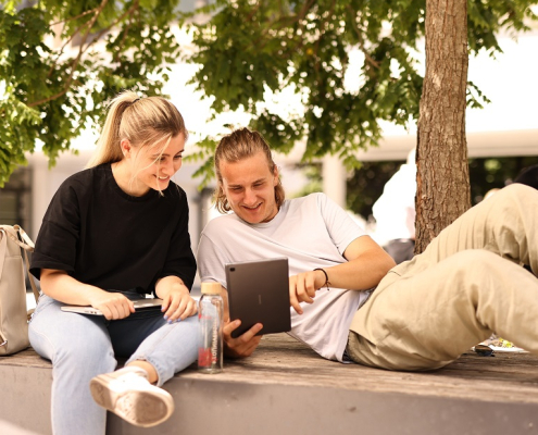 Zwei Studierende sitzen mit Touchpad und Laptop auf einer steinernen Sitzgelegenheit unter einem Baum.
