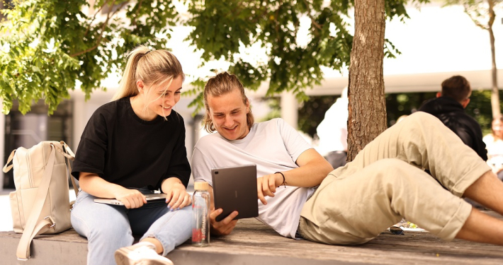 Zwei Studierende sitzen mit Touchpad und Laptop auf einer steinernen Sitzgelegenheit unter einem Baum.