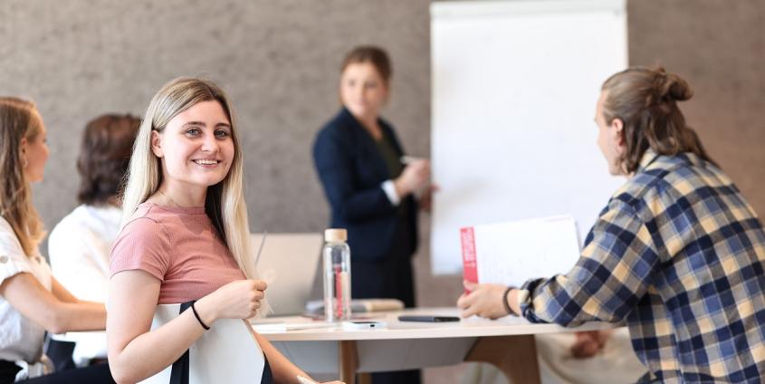 Eine Gruppe von vier Studierenden der Universität Klagenfurt sitzt an einem runden Tisch. Drei von ihnen schauen auf einen Flipchart im Hintergrund, auf den die den Kurs leitende Person gerade etwas aufzeichnet. Eine der Personen dreht sich um und schaut in die Kamera.