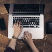 Eine Person tippt auf einem Laptop. Daneben liegen ein Smartphone, eine Geldbörse und eine Kamera auf einem Holztisch.